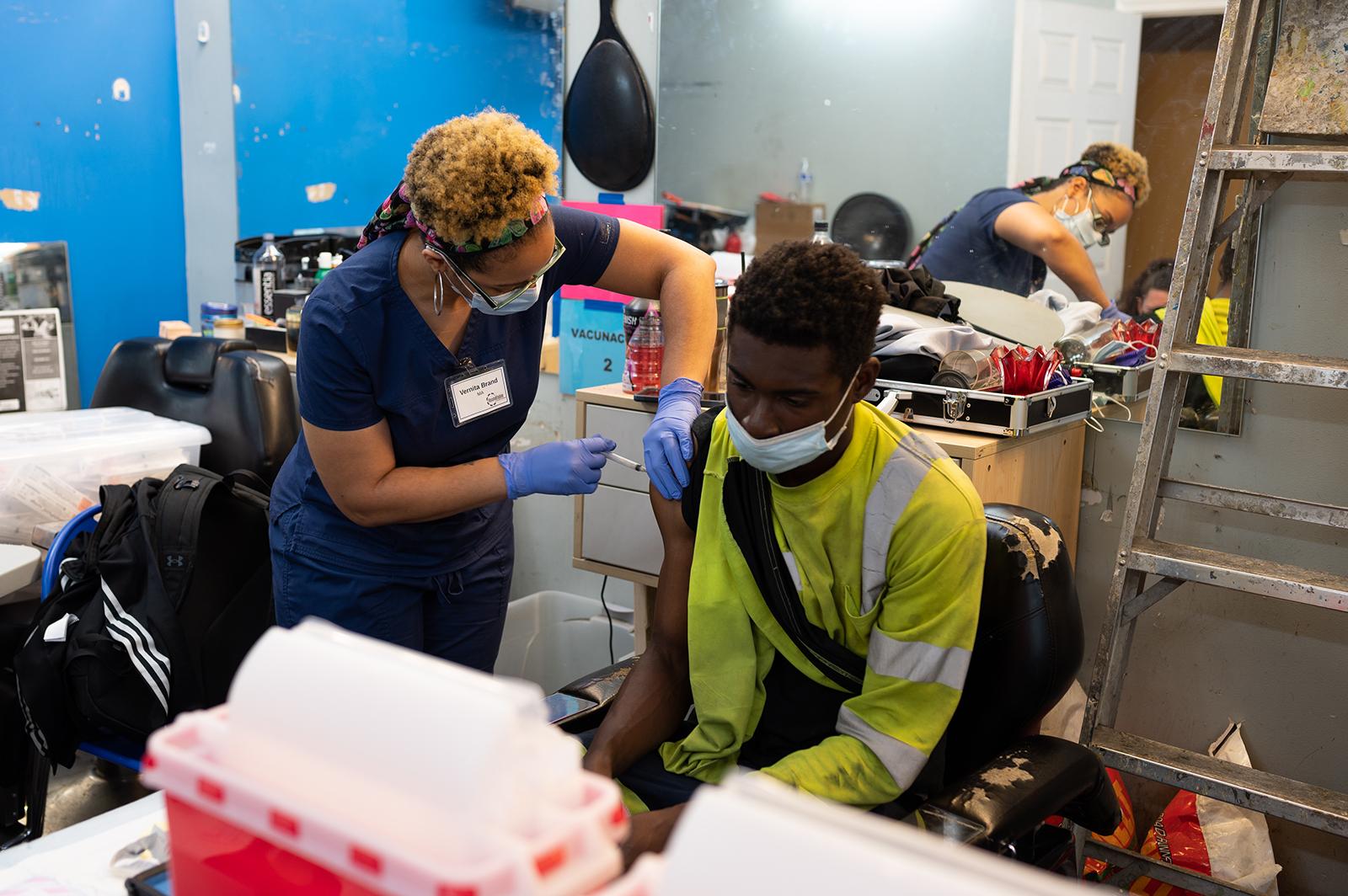 Black female nurse standing up and giving COVID vaccine in the arm of a young Black man who is seated and wearing construction worker clothing.