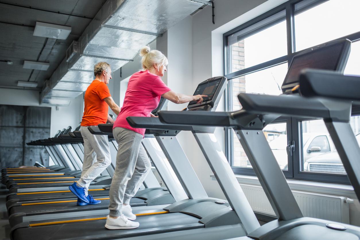 Two white people, one male and one female, walk on treadmills indoors. 