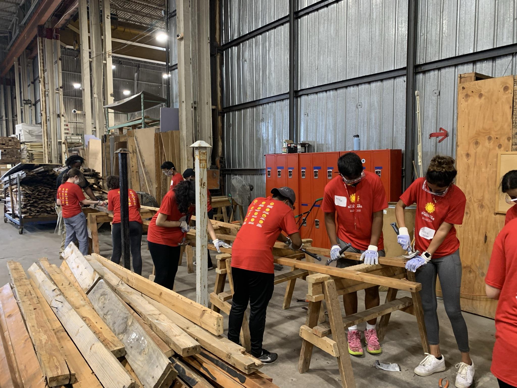 Global health scholars in red shirts stand a long a work bench, removing nails from planks of wood with hammers.