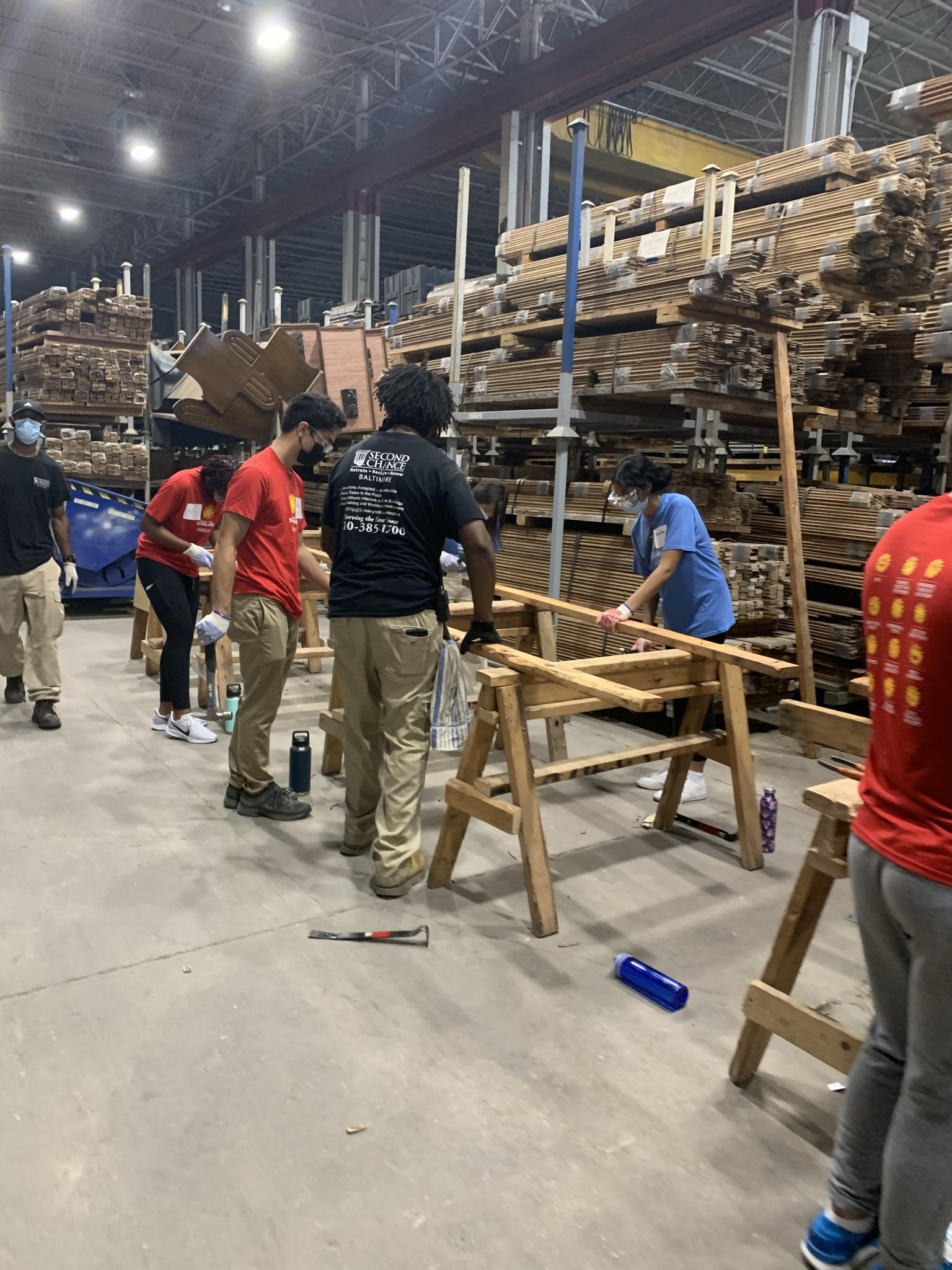 Students work in the Second Chance warehouse with wooden planks. Someone wearing a black Second Chance shirt is guiding them.