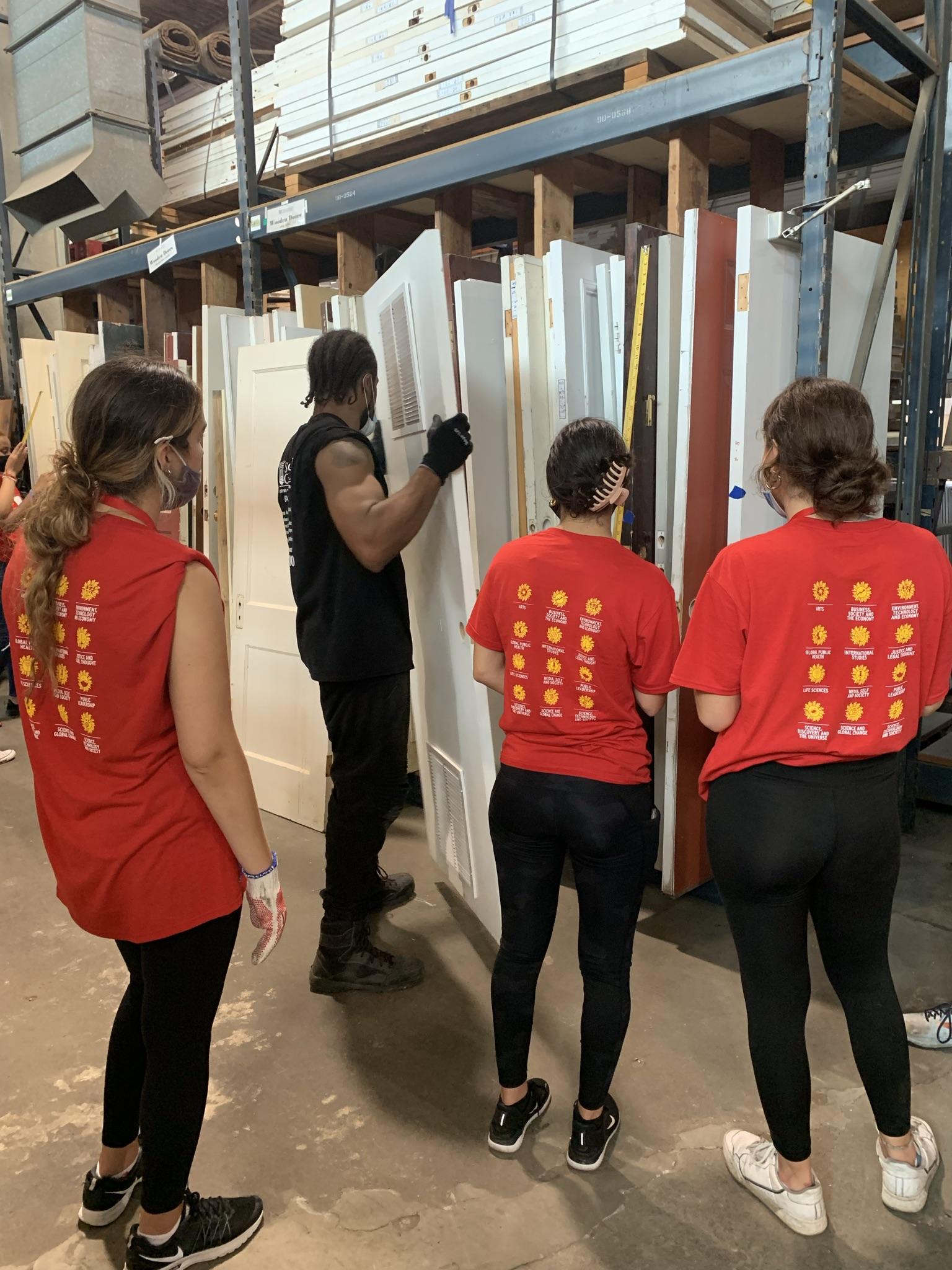 Three global health scholars look on as a man in a Second Chance shirt carries a white wooden door. There is a rack of doors with different styles in front of them.