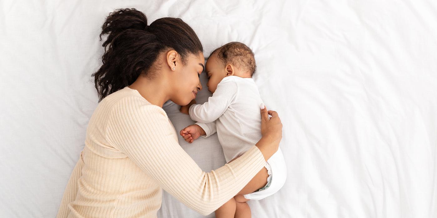 Young African American mom and infant baby laying together face to face on white bed