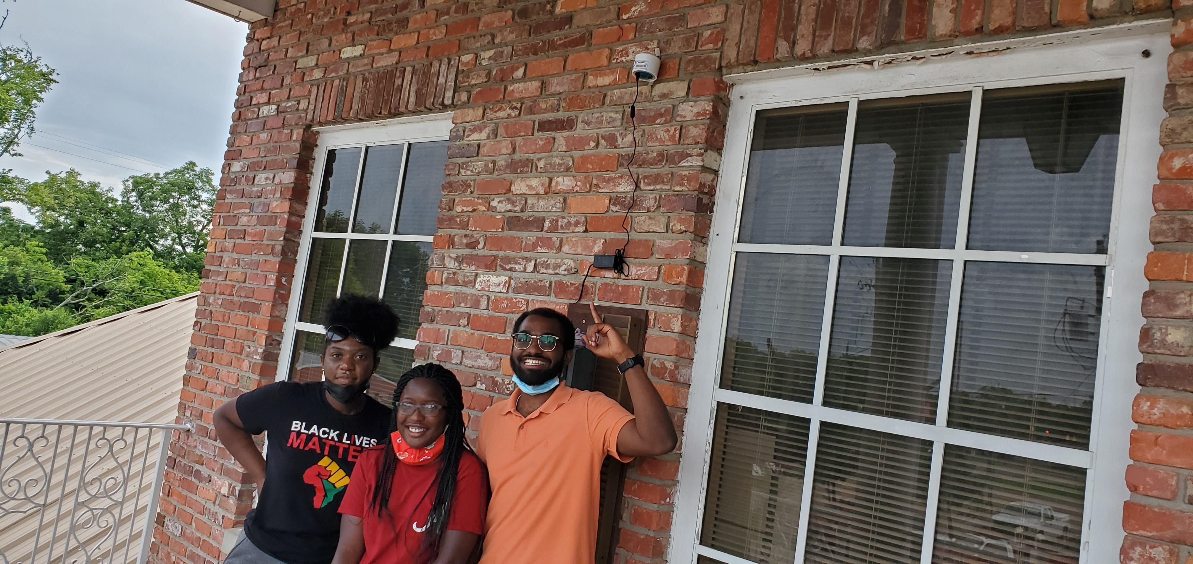 Doctoral student Jan-Michael Archer (on right) points to an air quality monitoring device he installed at a home in Cheverly, Maryland. Two women pose with him.