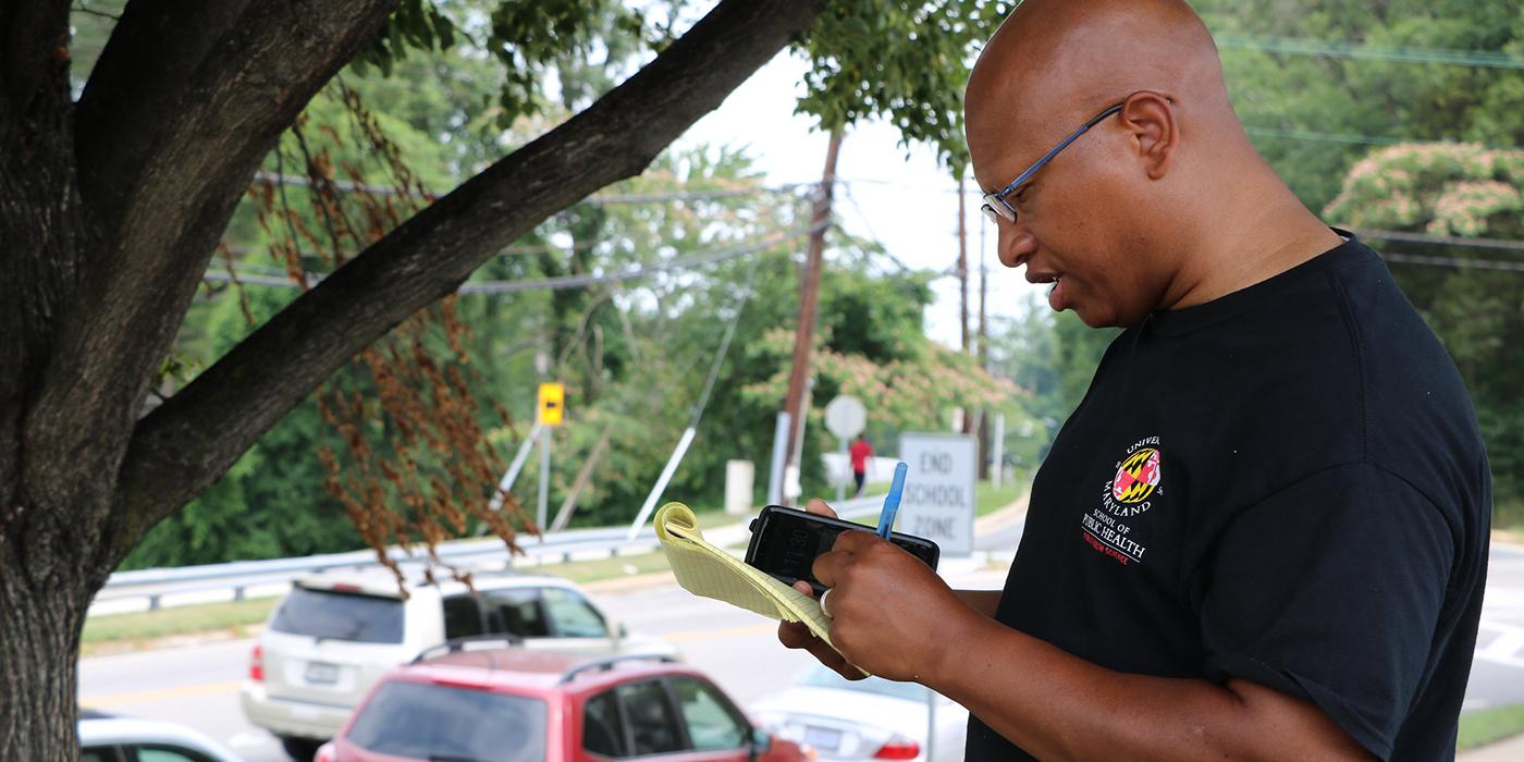 Sacoby Wilson, an African American man with glasses and a bald head writes on a small yellow notebook and holds his phone in his hand. He is outside monitoring air quality in Langley Park, Maryland and there are cars passing by.