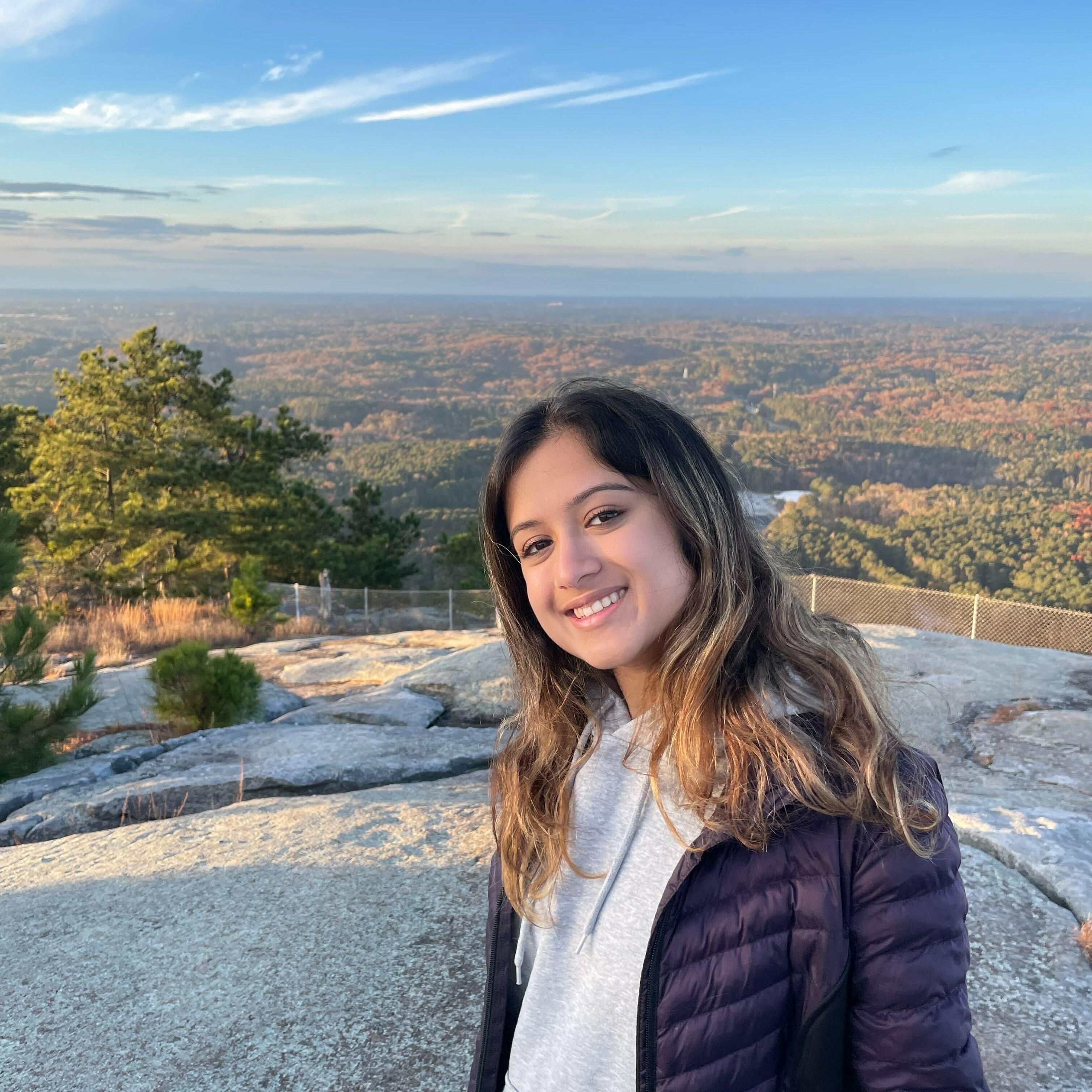 Esha Saxen smiles at the camera with a beautiful view of trees and blue skies behind her.