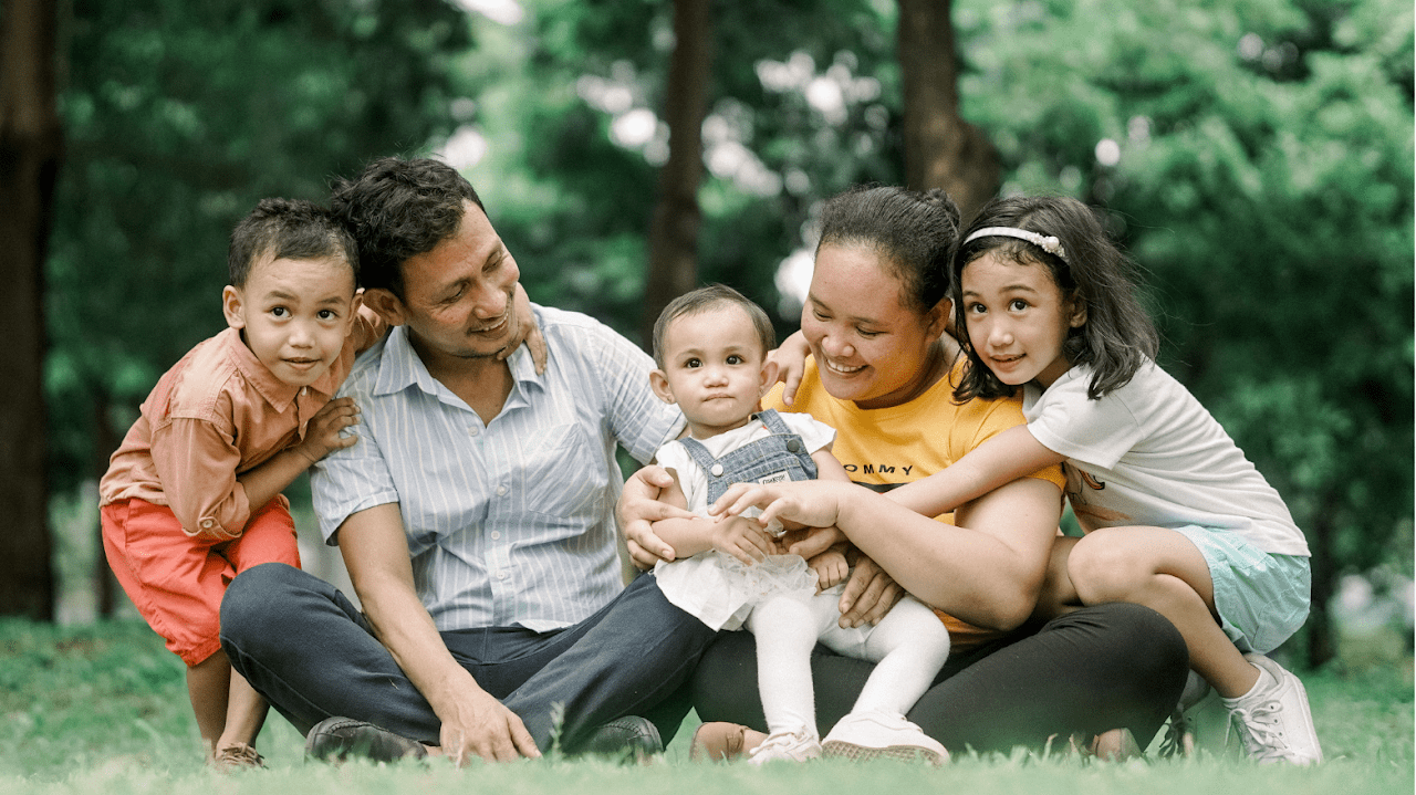 Hispanic looking parents sitting in the grass with three small children sitting and hanging on them