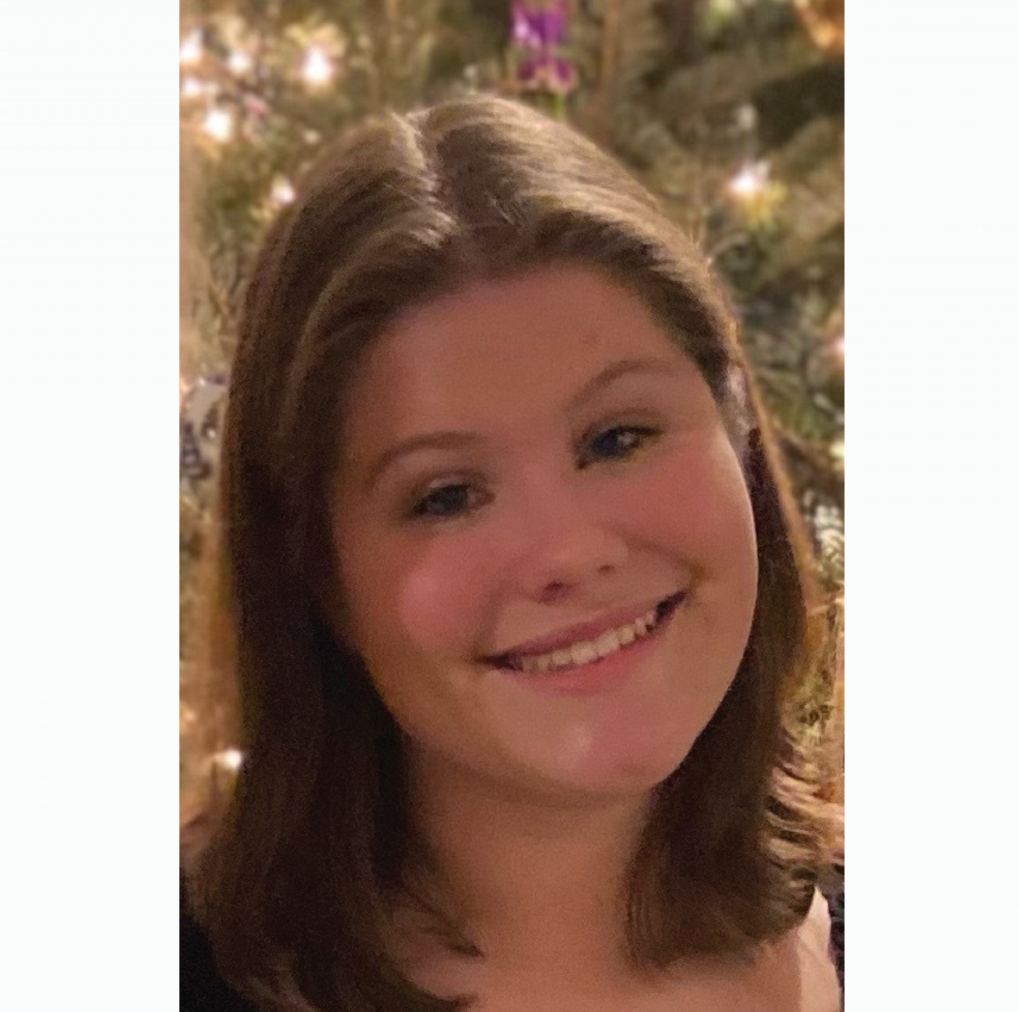 Mya smiles at the camera while standing in front of a christmas tree with sparkling lights.