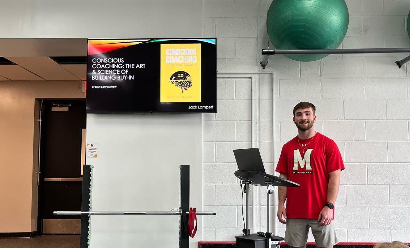 Man wearing red shirt with M logo stands under a TV