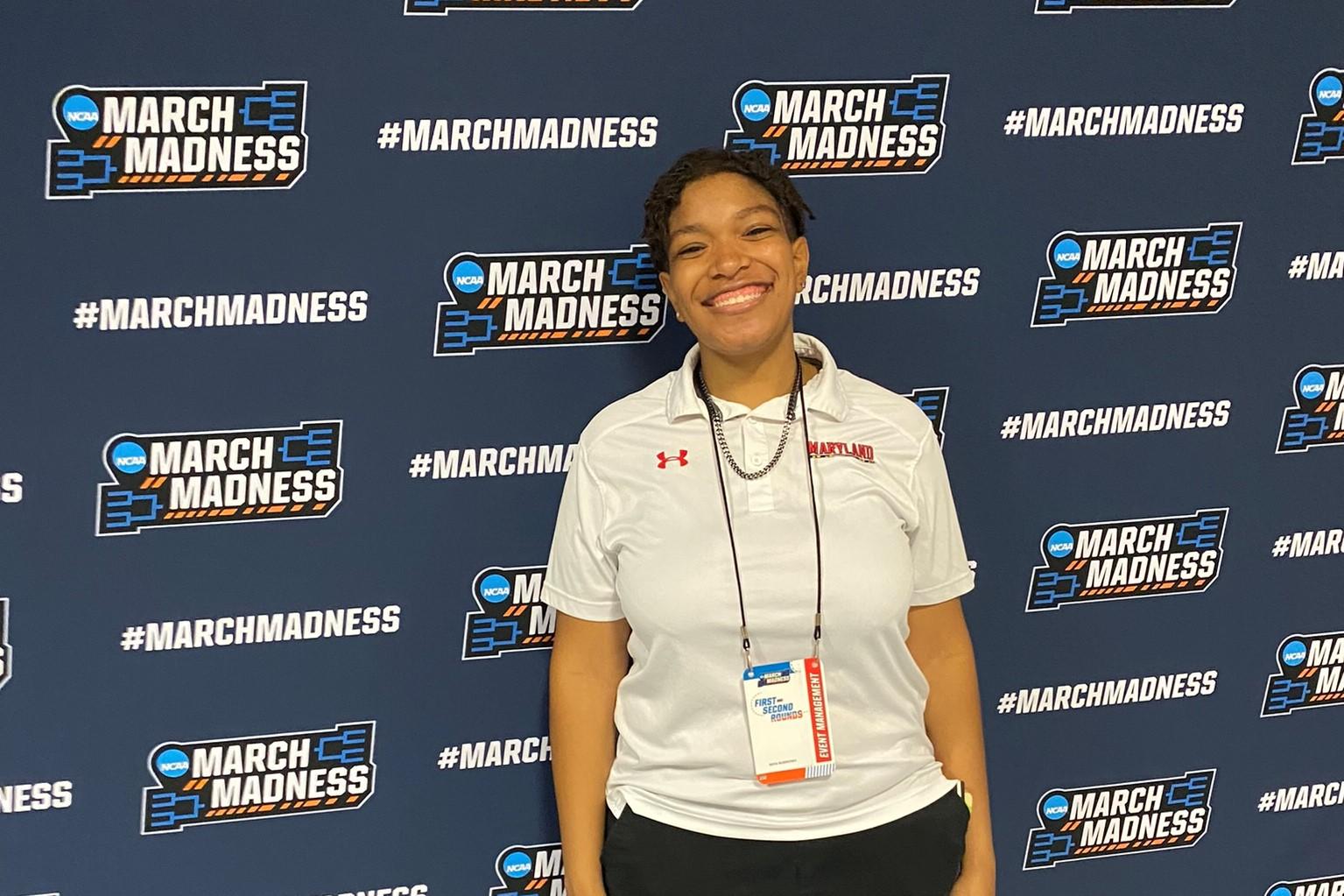 Student wearing gray polo and lanyard around their neck stands in front of a blue March Madness banner.