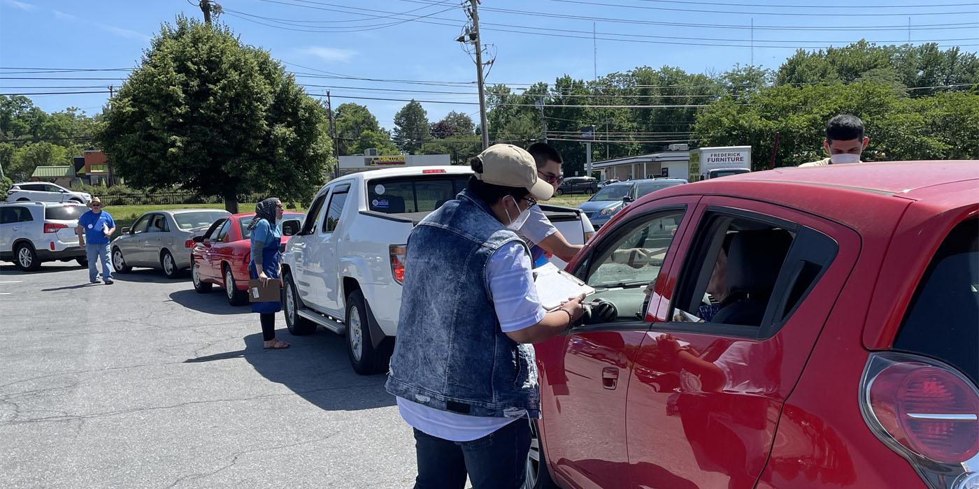 In the foreground, a community health worker with a clip board leans close to a car window waiting in a line of cars. In the background, other workers approach other cars.