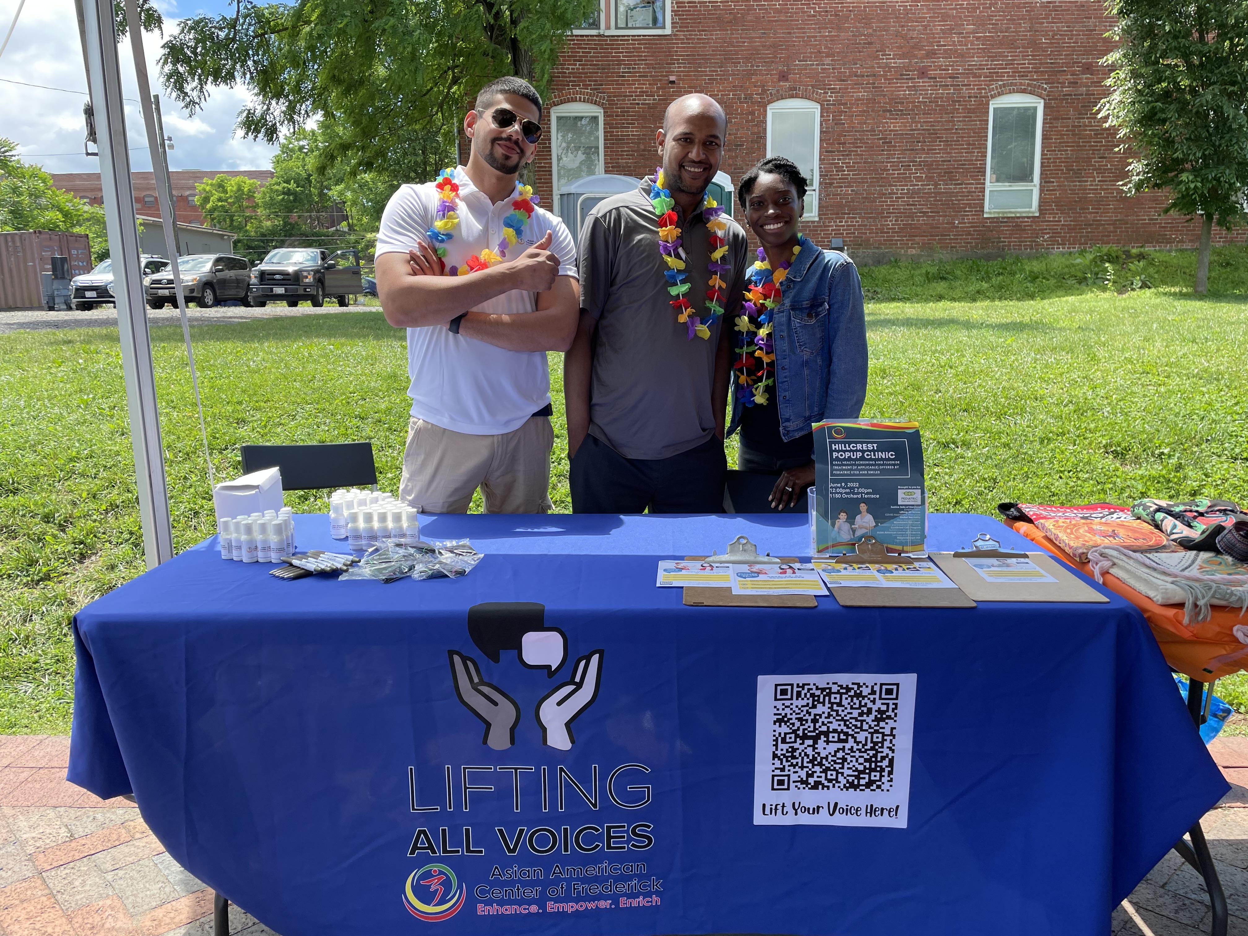 Three people stand smiling outside behind a folding table with a table cloth that reads "Lifting All Voices: Asian American Center of Frederick"