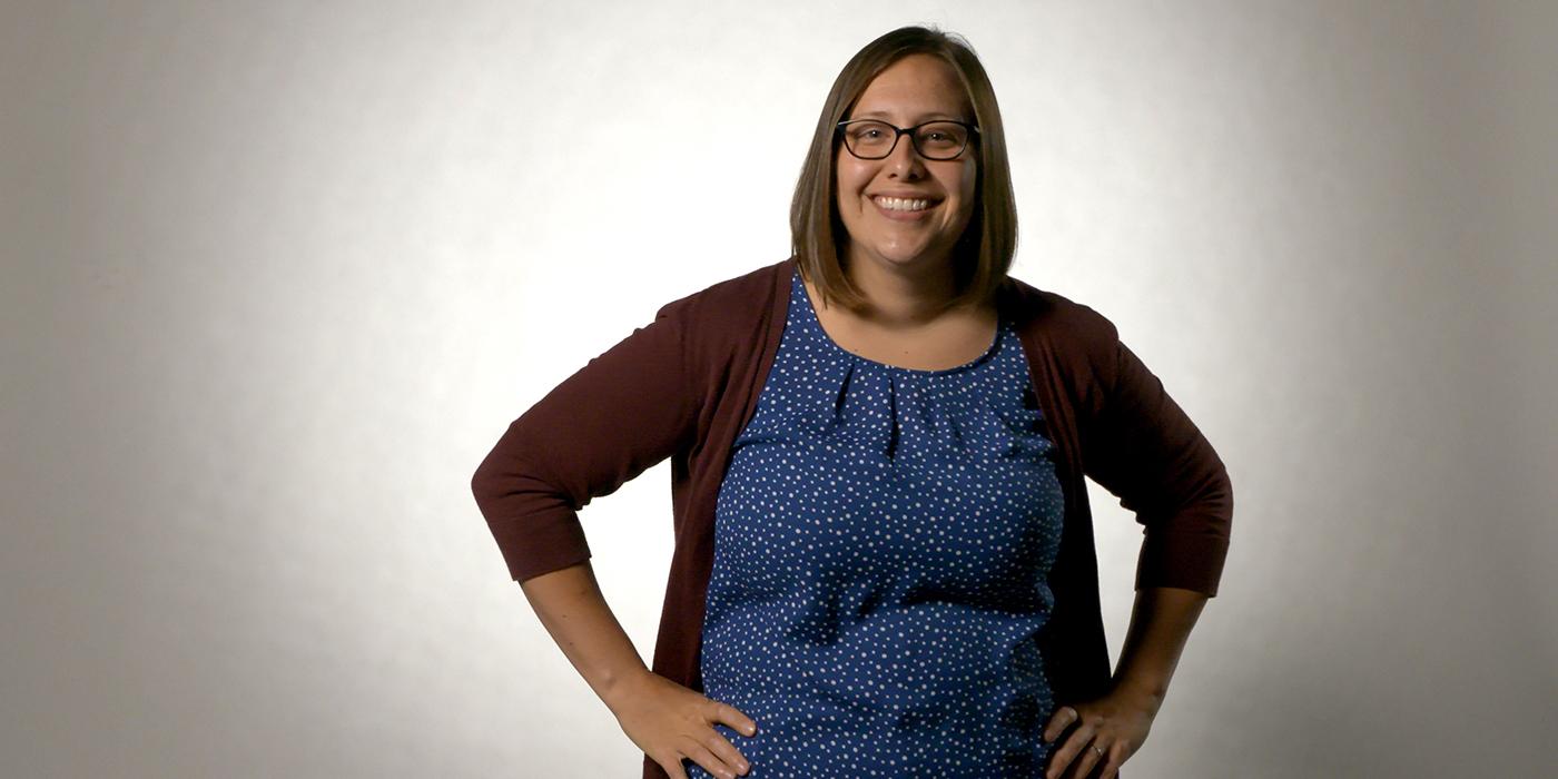 Jessica Fish, a white cis gendered woman with shoulder length brown hair wearing glasses, stands with her hands on her hips
