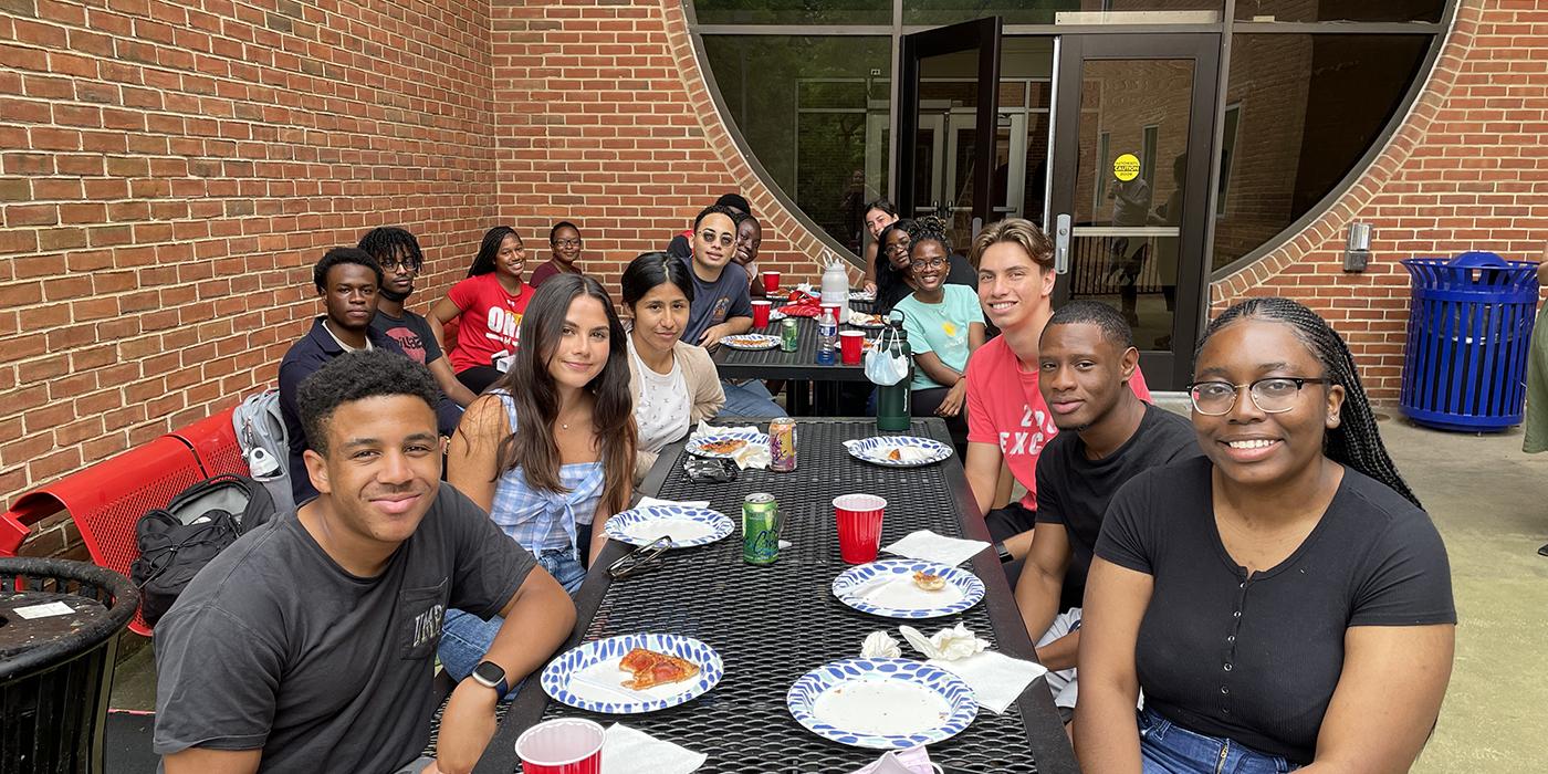 A group of African American, Latino and white UMD college students sit outside at a picnic table with pizza on paper plates. 