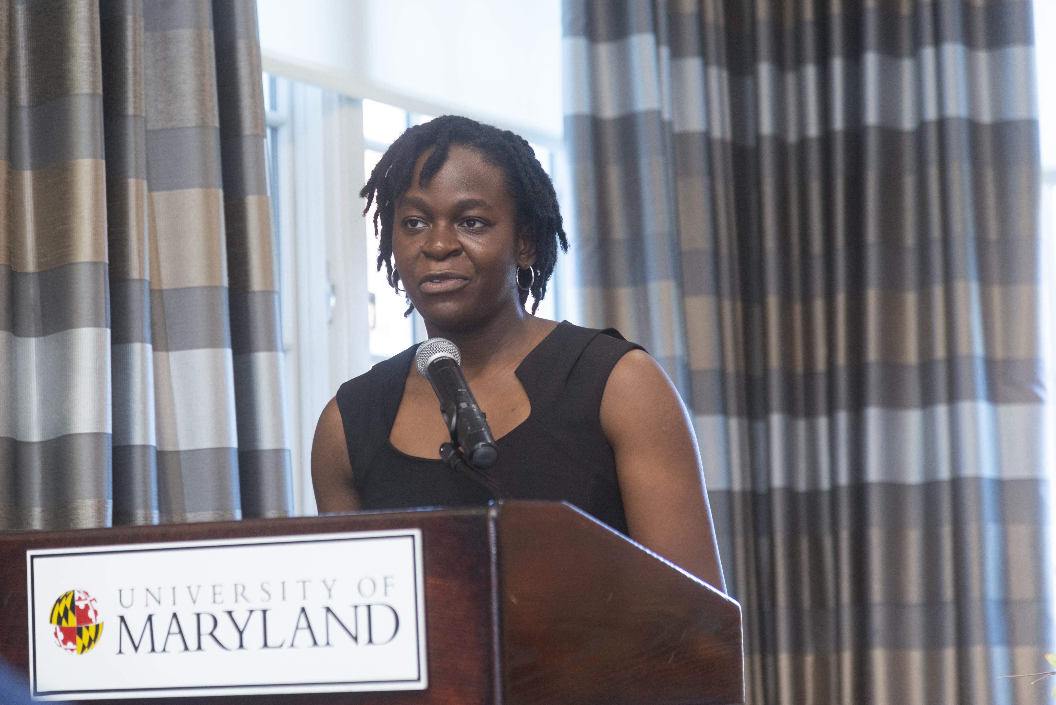 Deborah Omotoso, wearing a black sleeveless dress, speaks at a podium with the University of Maryland name on it.