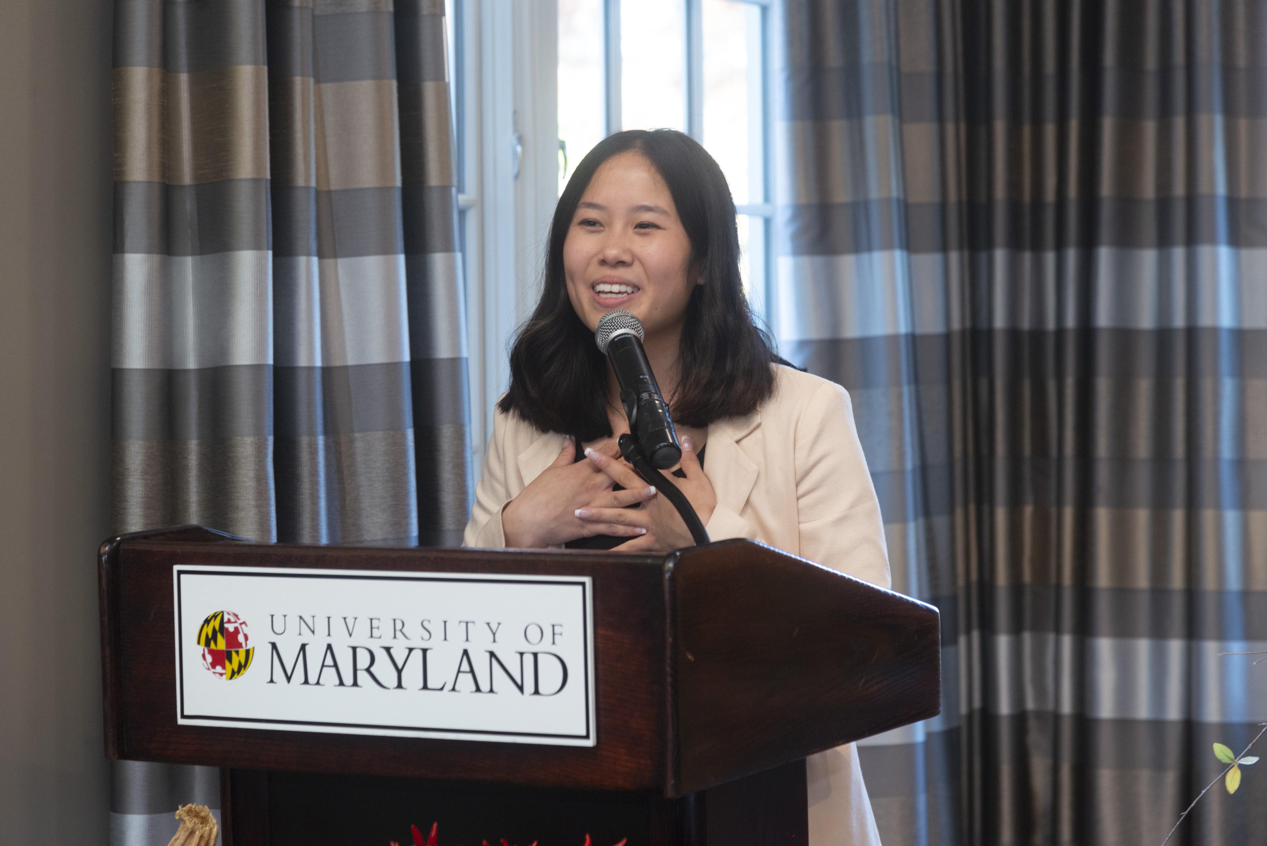 Katelyn Wang, wearing cream blazer and black shirt, speaks behind a podium with the University of Maryland name on it.