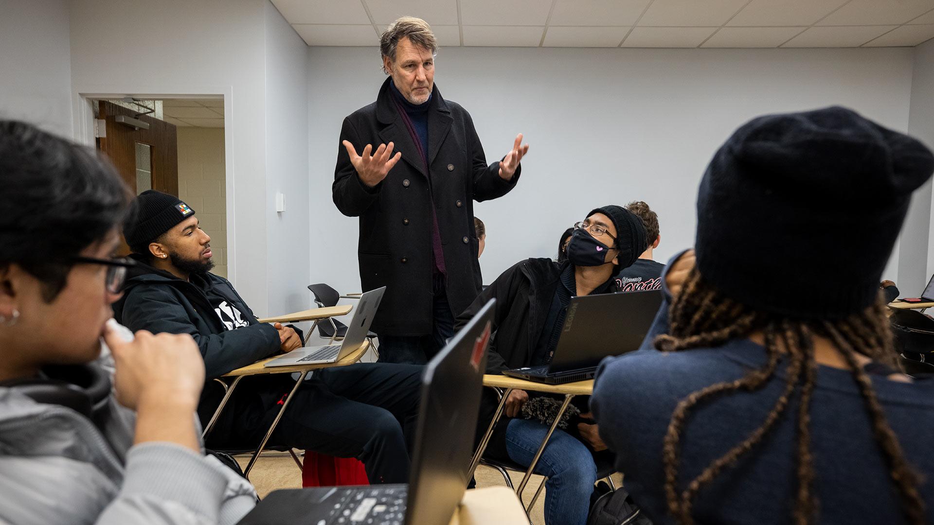 Male professor wearing black jacket stands with arms outstretched in the middle of a group of seated students.