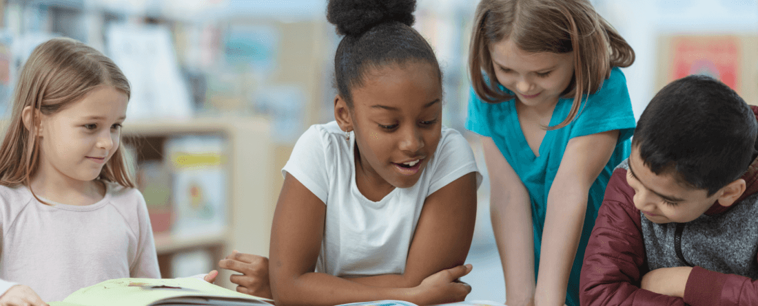 Four children crowd together around a book on a table.