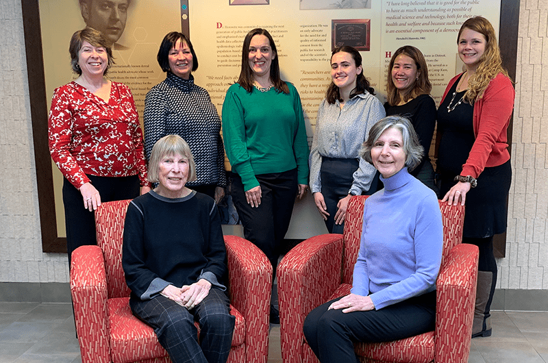 Group photo of Horowitz Center staff and stake holders smiling in the school of public health in front of the poster about Dr. Hershel Horowitz.