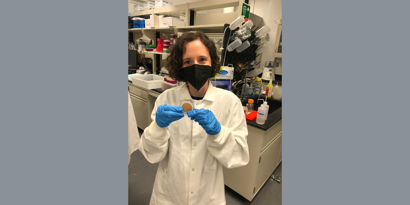 Woman wearing a black mask and white lab coat standing inside a science lab.