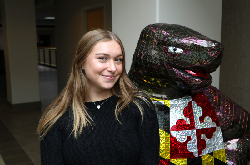 Young Caucasian woman with long brown hair smiling next to a Testudo statue.