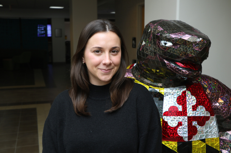 Young Caucasian woman with long dark brown hair smiling next to a Testudo statue.