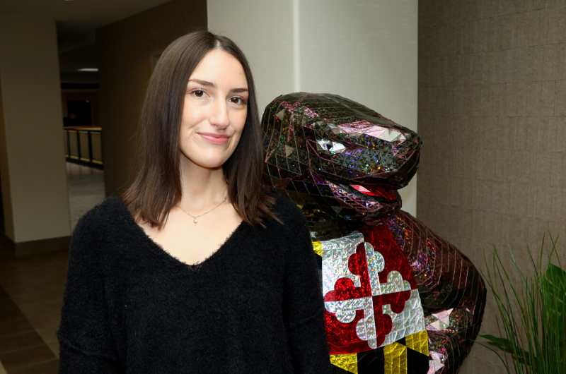 Young Caucasian woman with long dark brown hair smiling next to a Testudo statue.