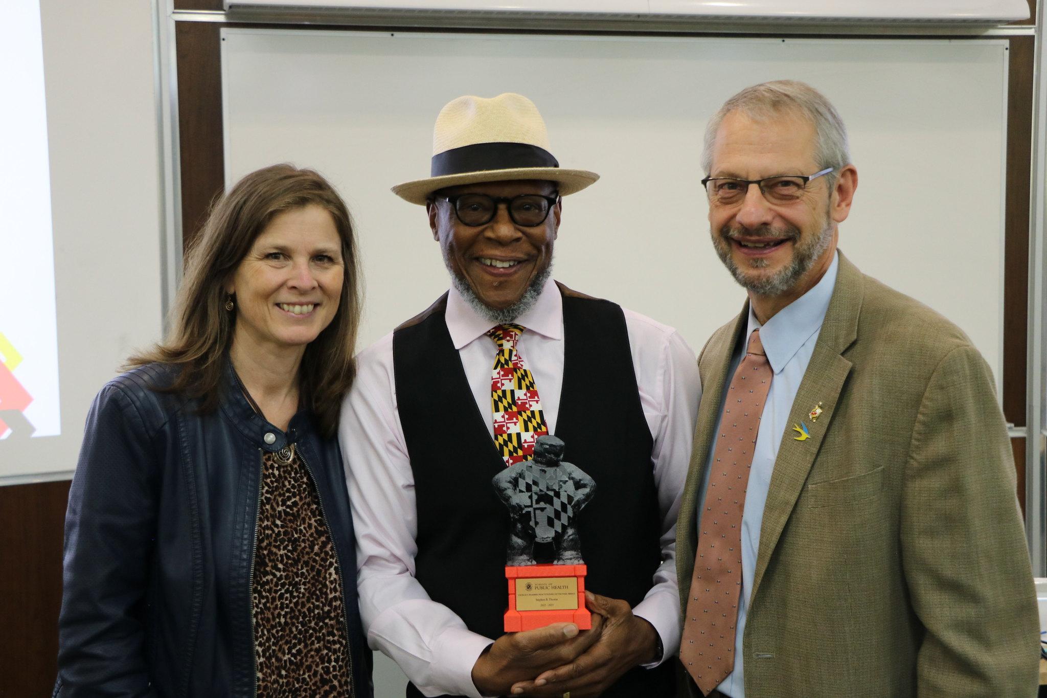 Man holding an award and wearing a hat stands in between a woman to the left and a man to the right.