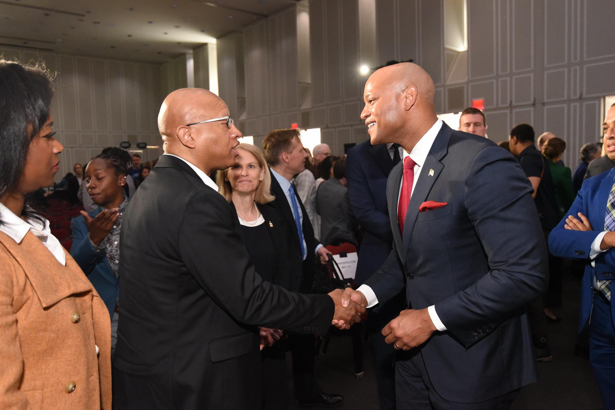 Two men shake hands in a conference room as others look on.