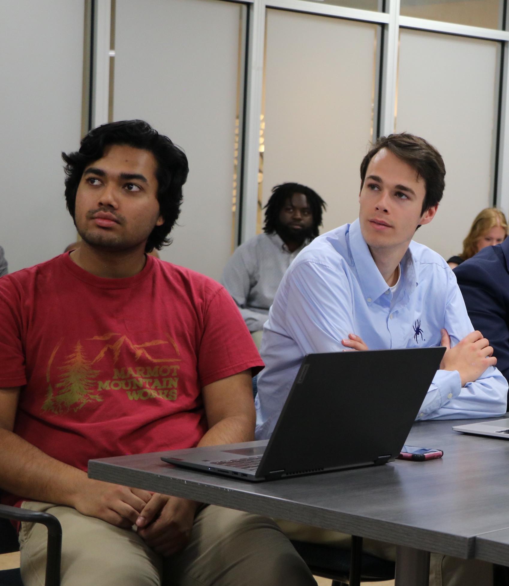 Two male students sit inside a classroom behind a laptop computer.