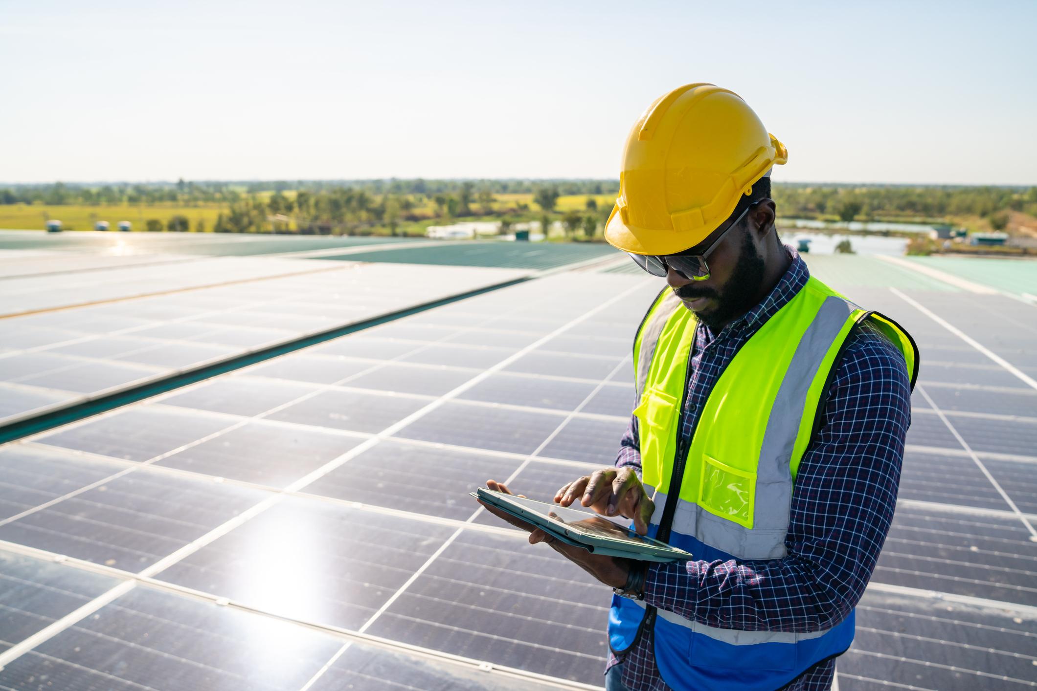 Man with a hard hat and reflector vest standing with solar panels looking at a work tablet