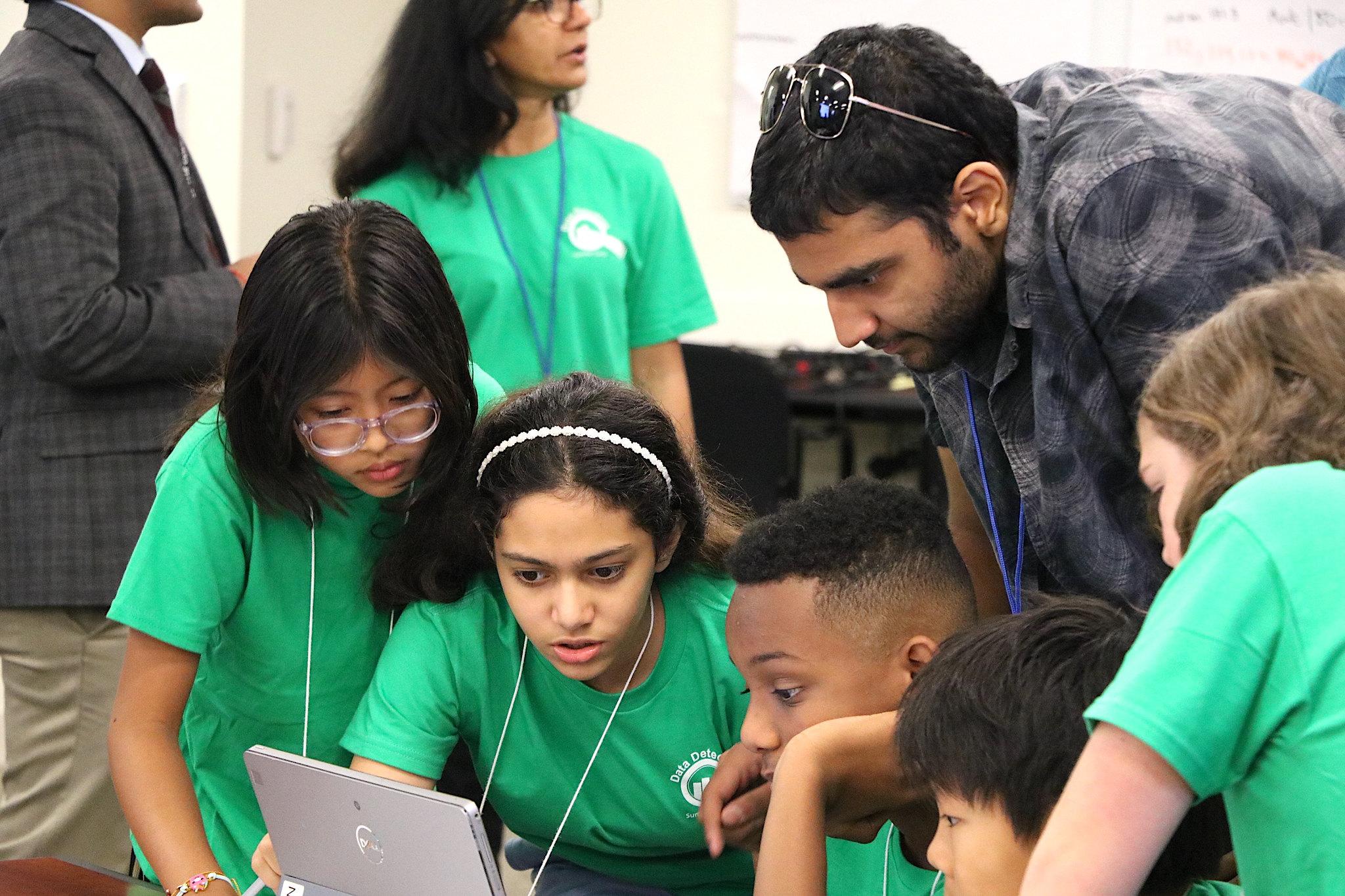 Students from the Data Detectives Camp wearing green t-shirts gather around a table while looking at a computer screen.