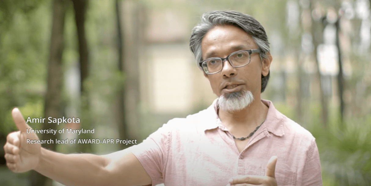 Professor wearing pink shirt reaches his arm out to the right, trees in the background.