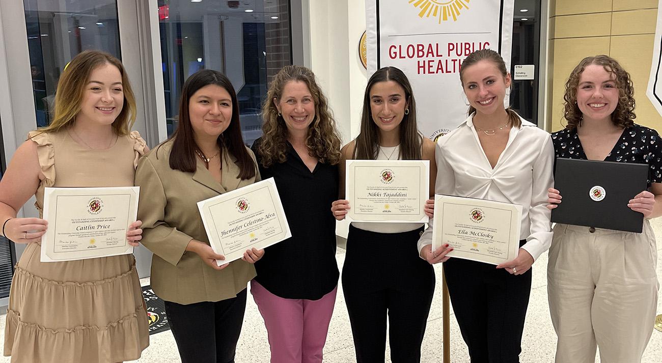 A group of five college students hold awards certificates, and a faculty member stands in the middle of the group smiling.