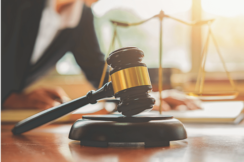 A gavel rests on a desk in front of scales of justice; a person is in the background