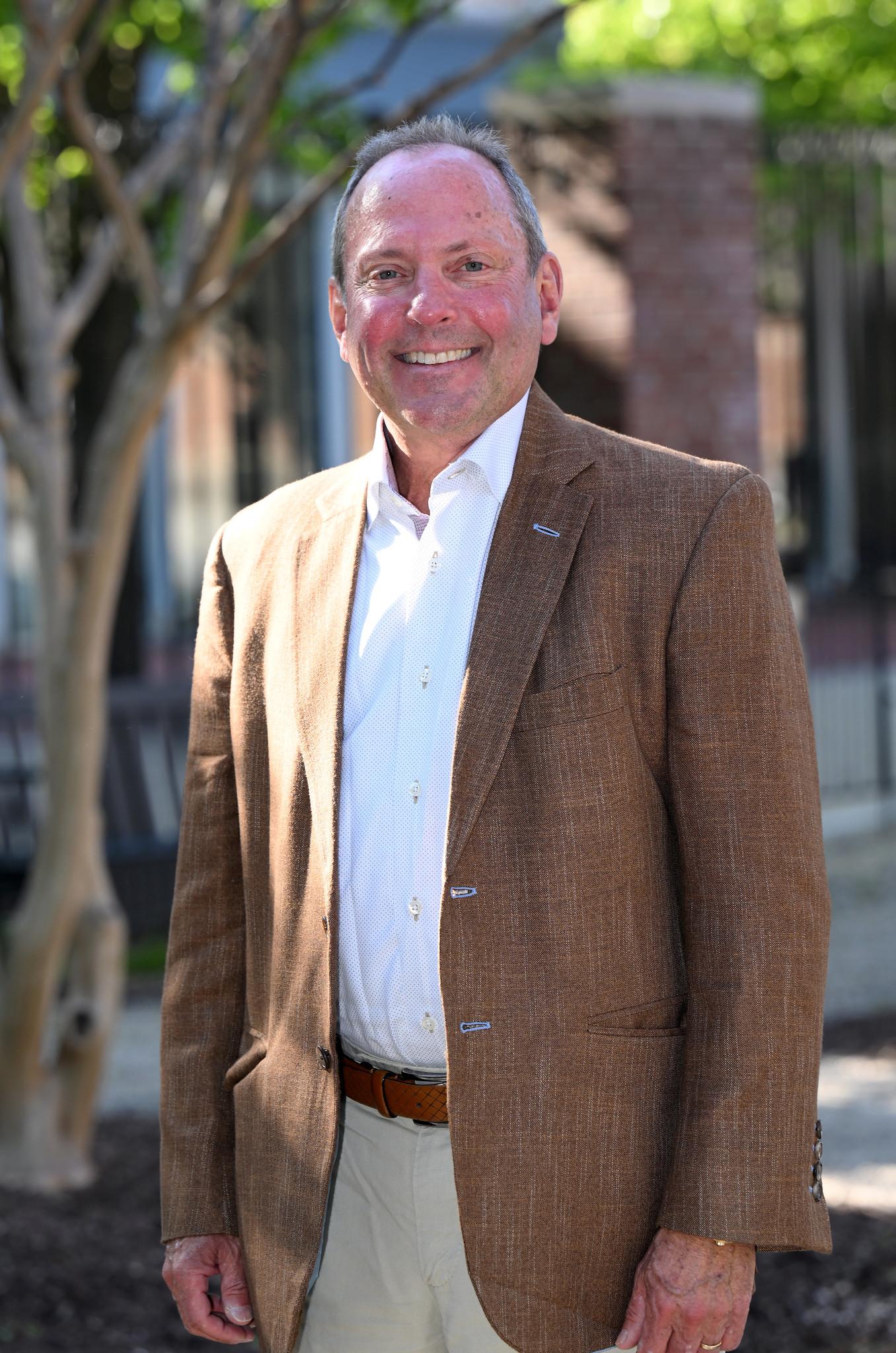 Alumni white man in a brown and beige suite stands outside in front of trees