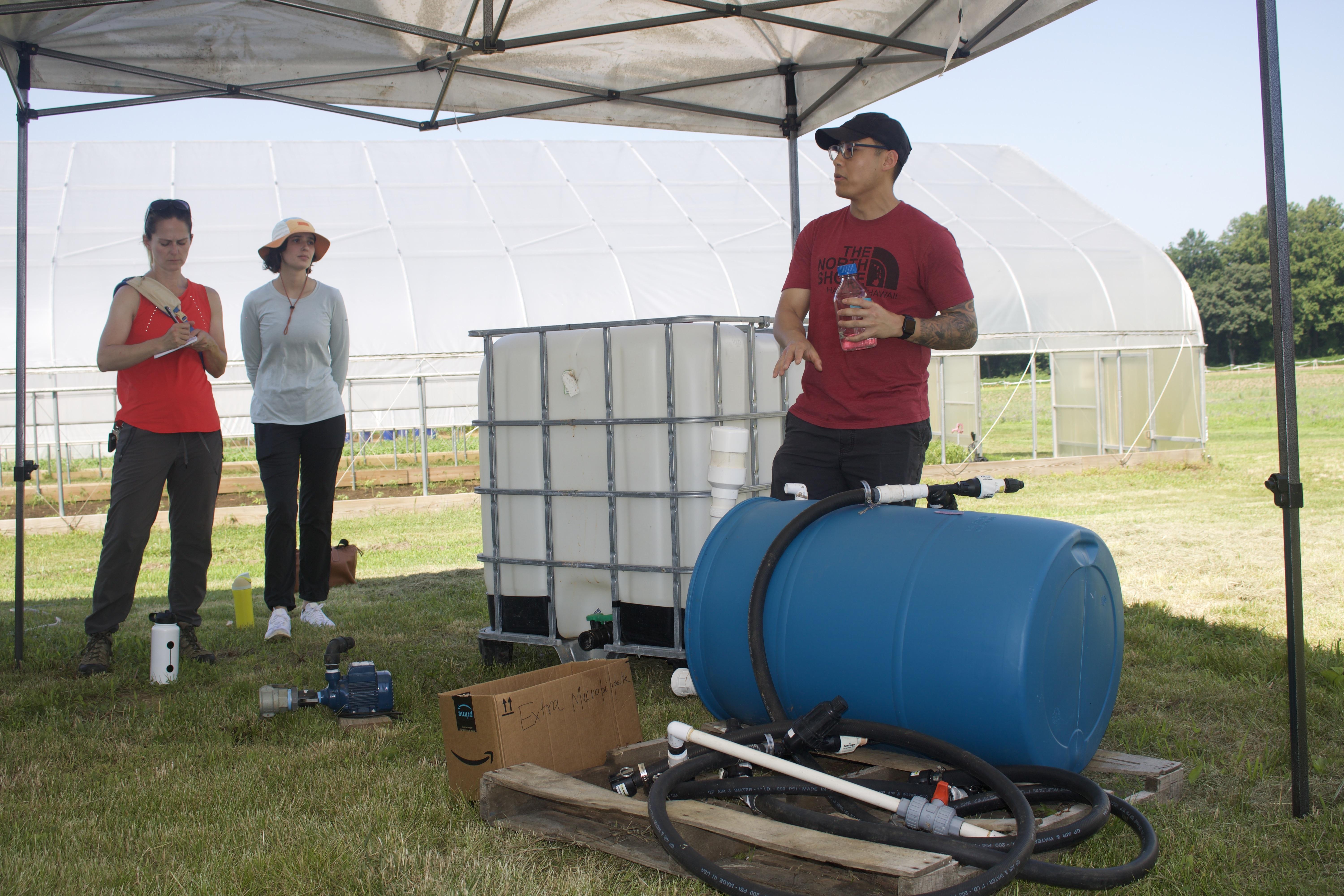 scientist in red tshirt stands in front of rainwater harvesting equipment and talks to a group