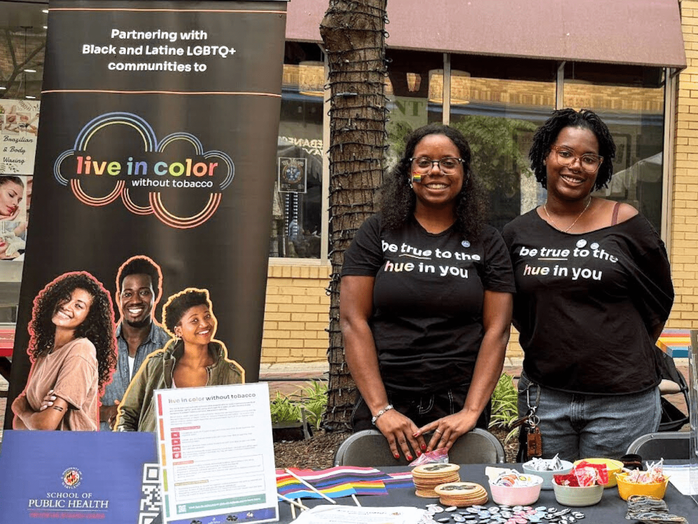 two black women wearing tshirts that read Stay True To Your Hue stand by a banner with LIVE IN COLOR on it