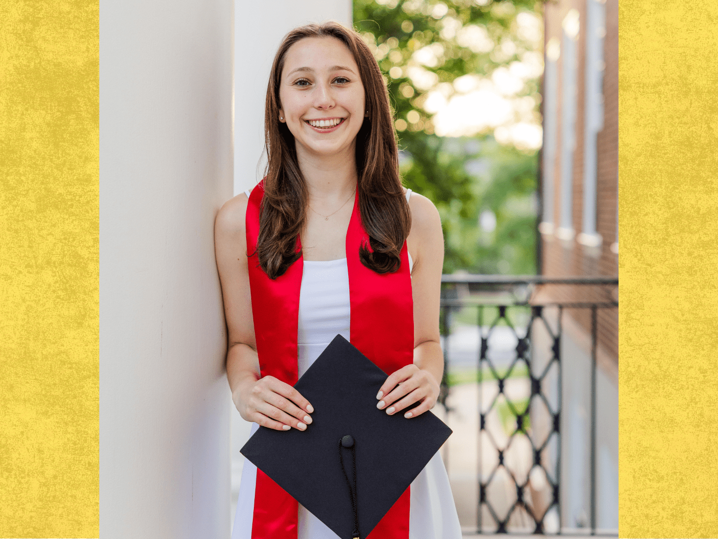 Student in white dress and red graduation scarf, holds black graduation hat, smiles to camera