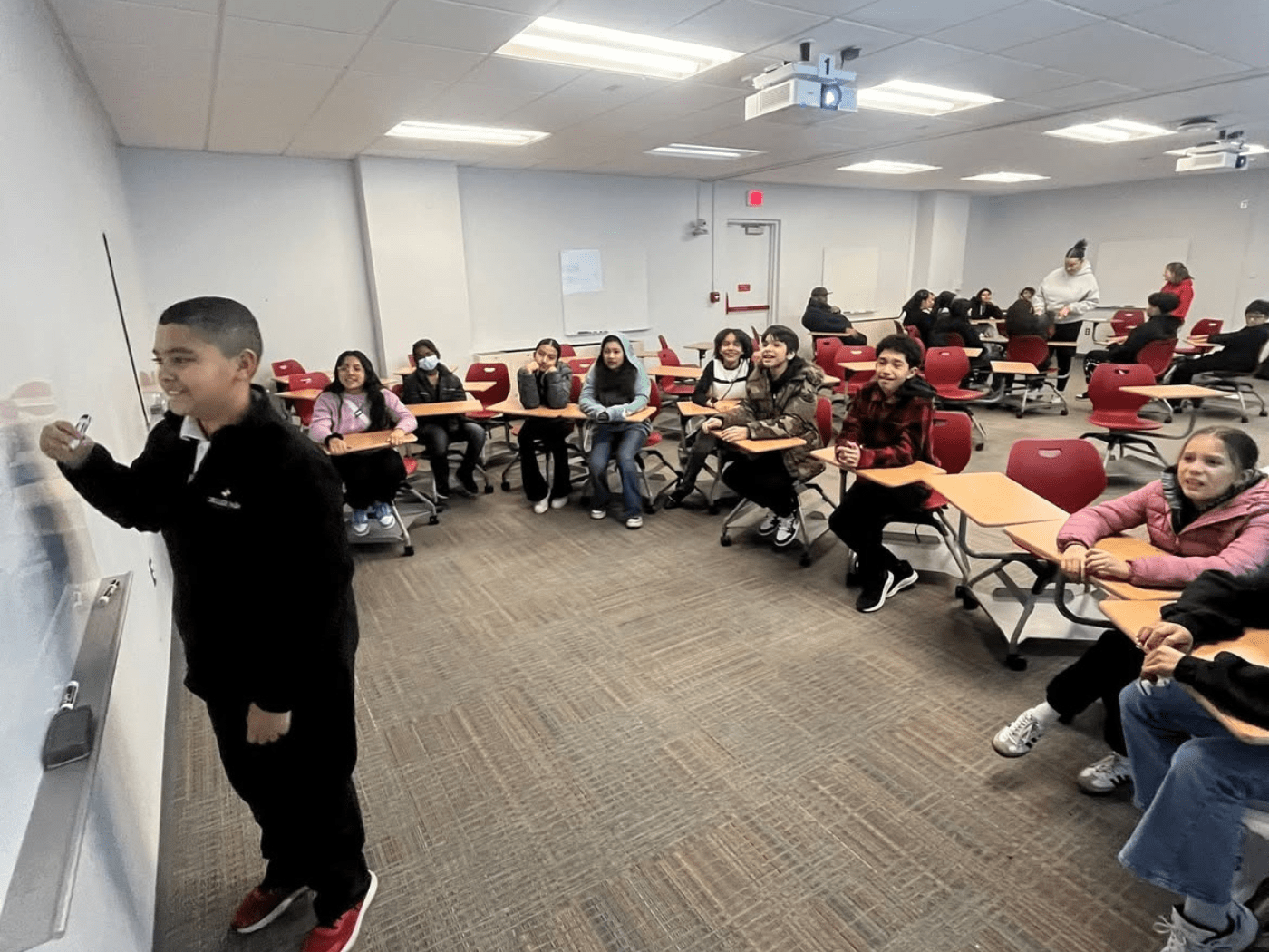 young boy student writes on white board with semi circle of fellow students watching him
