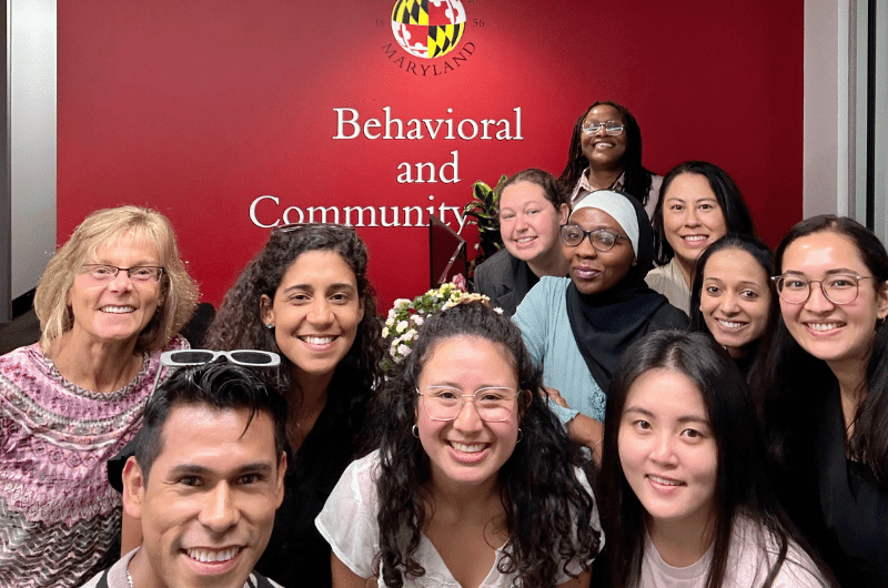 Group of eleven smiling people in front of a red University of Maryland wall that reads “Behavioral and Community Health.