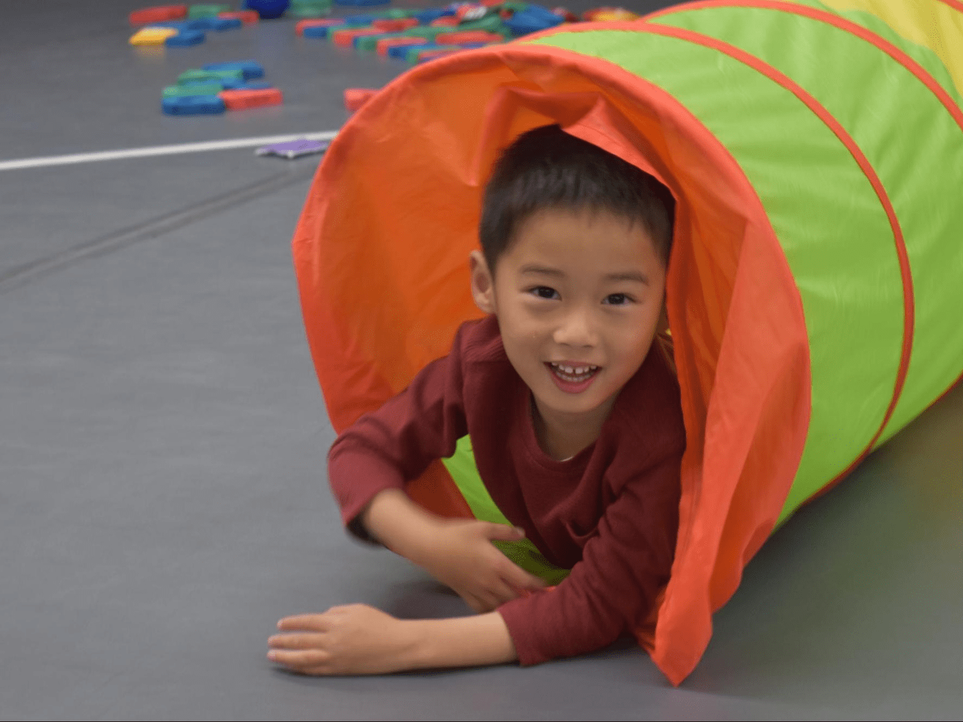 young asian boy in red longsleeve shirt smiles, as he crawls out of a mesh tunnel in the gym