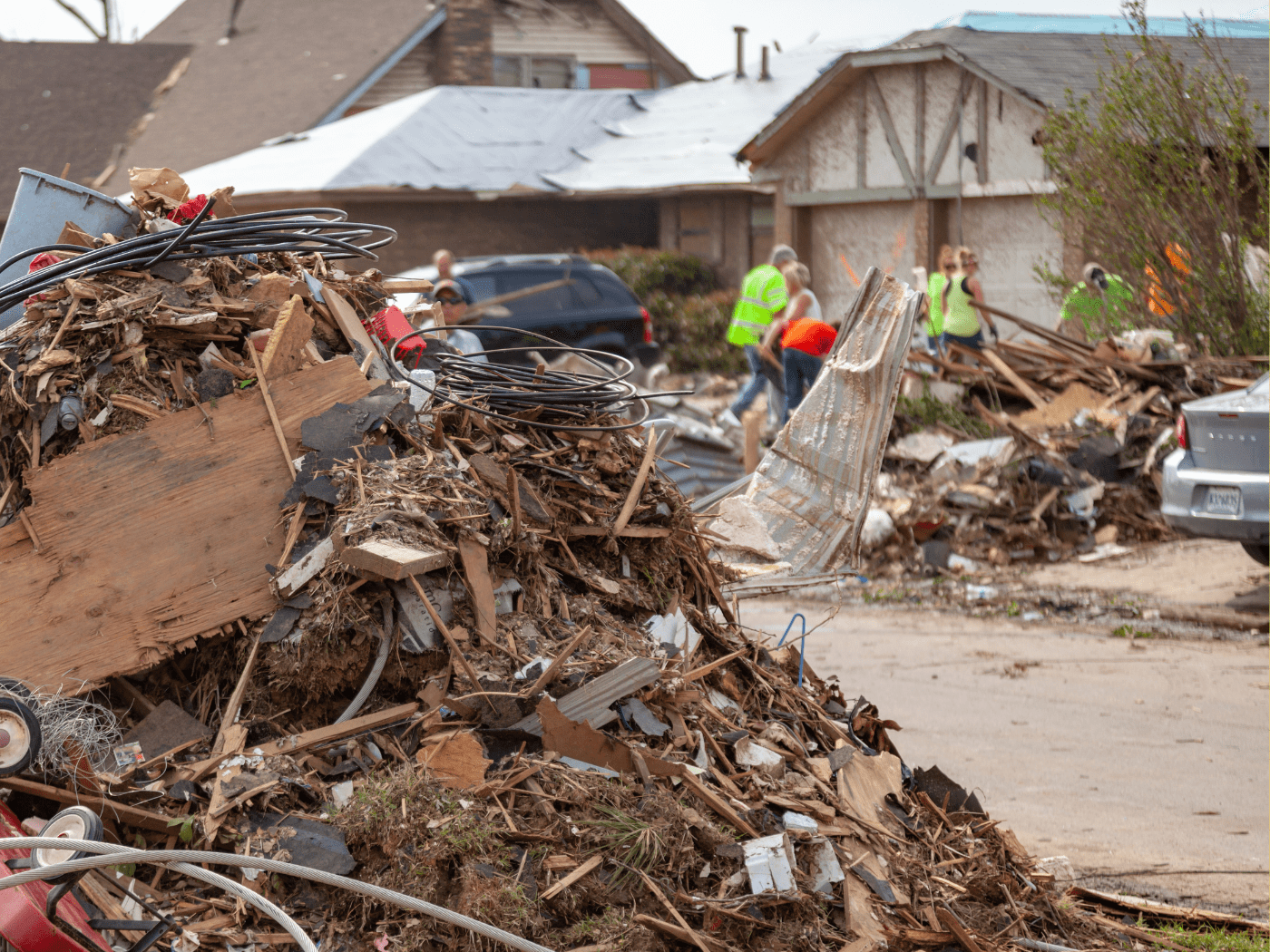 debris of destroyed house is piled up to the left, in background people in neon safety vests clean up