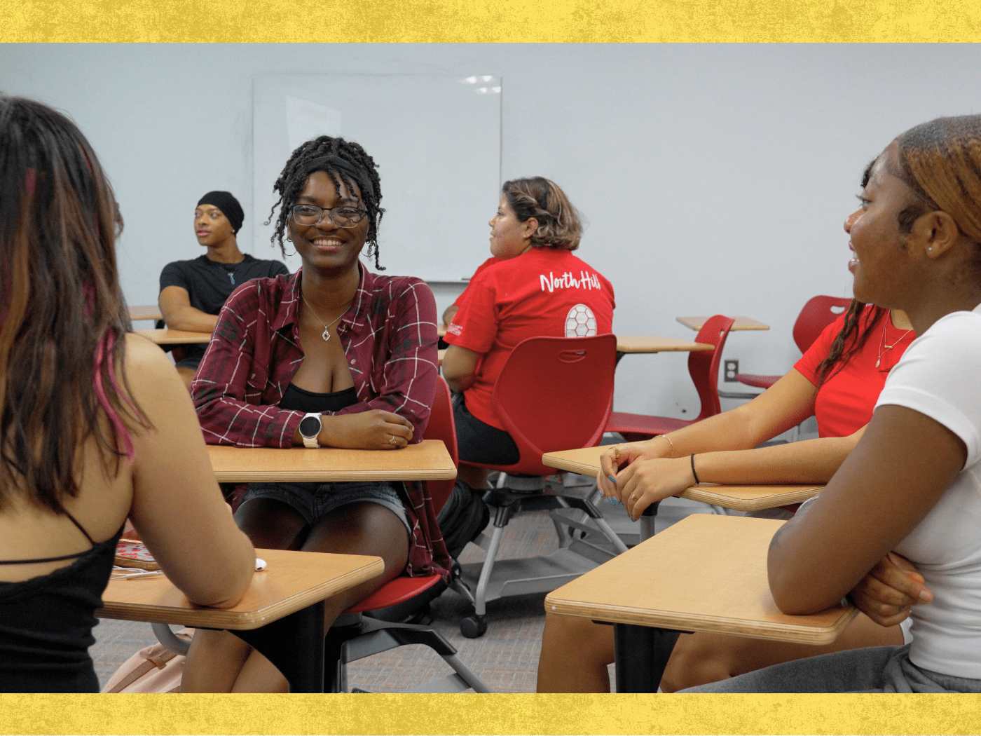 students sit at single desks in a small group talking in a classroom space, with another student group visible in the background