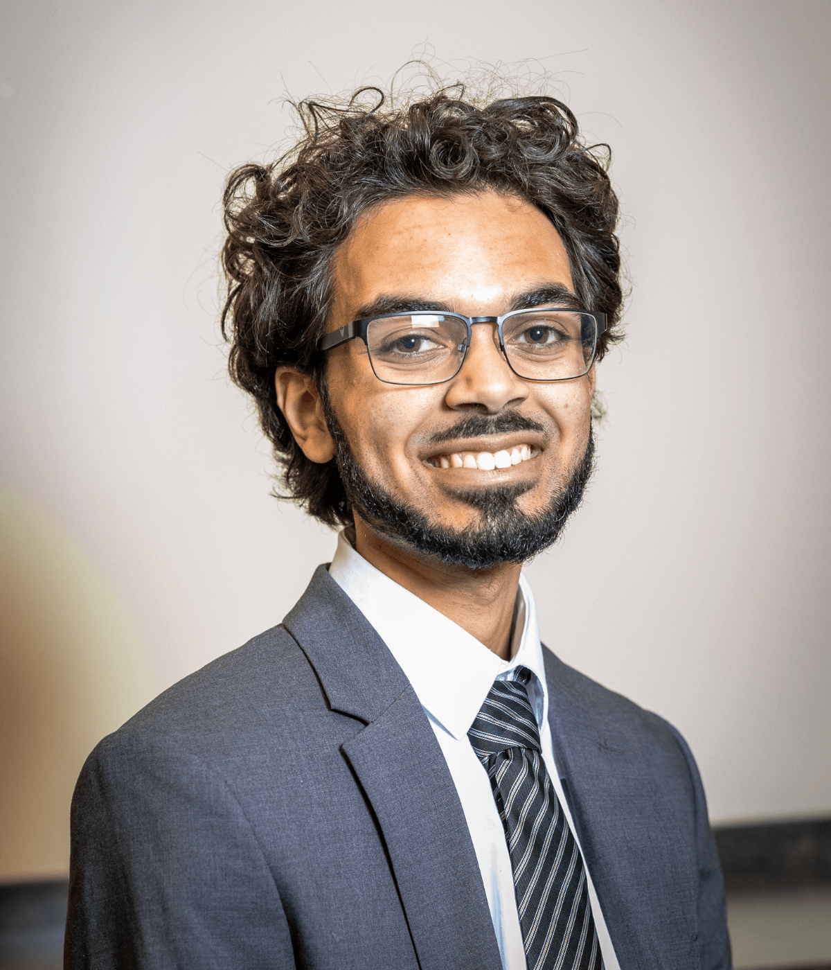 Portrait of a young man with curly hair, glasses, and a short beard, smiling at the camera while wearing a gray suit, white shirt, and striped tie against a plain light-colored background.