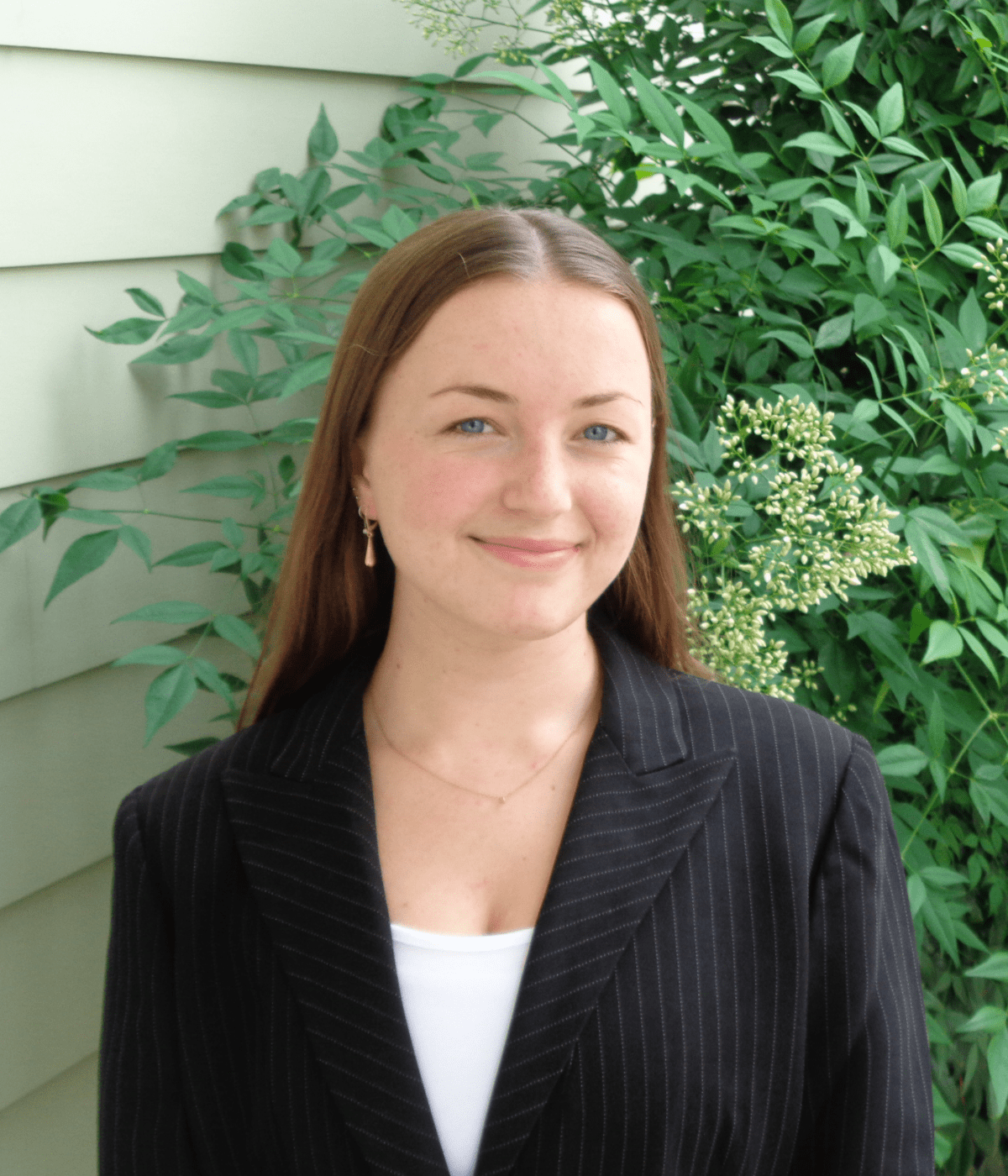 Portrait of a young woman with long straight hair wearing a black pinstripe blazer and white top, smiling at the camera while standing outdoors in front of leafy green plants and light-colored siding.