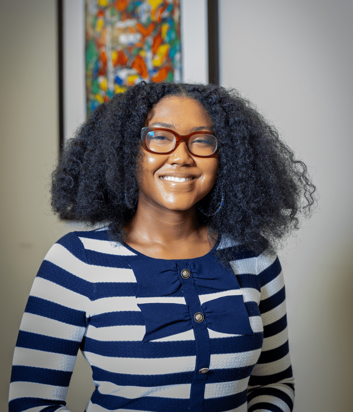 Portrait of a young woman with voluminous curly hair and glasses, smiling at the camera while wearing a navy-and-white striped top with bow accents, with a colorful abstract painting blurred in the background.