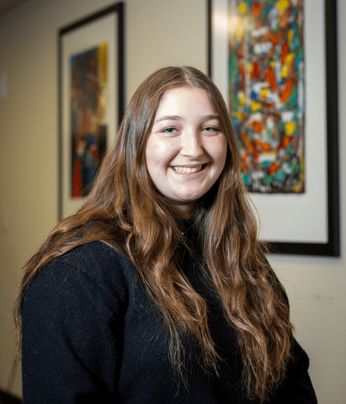 Portrait of a young woman with long straight hair wearing a black pinstripe blazer and white top, smiling at the camera while standing outdoors in front of leafy green plants and light-colored siding.