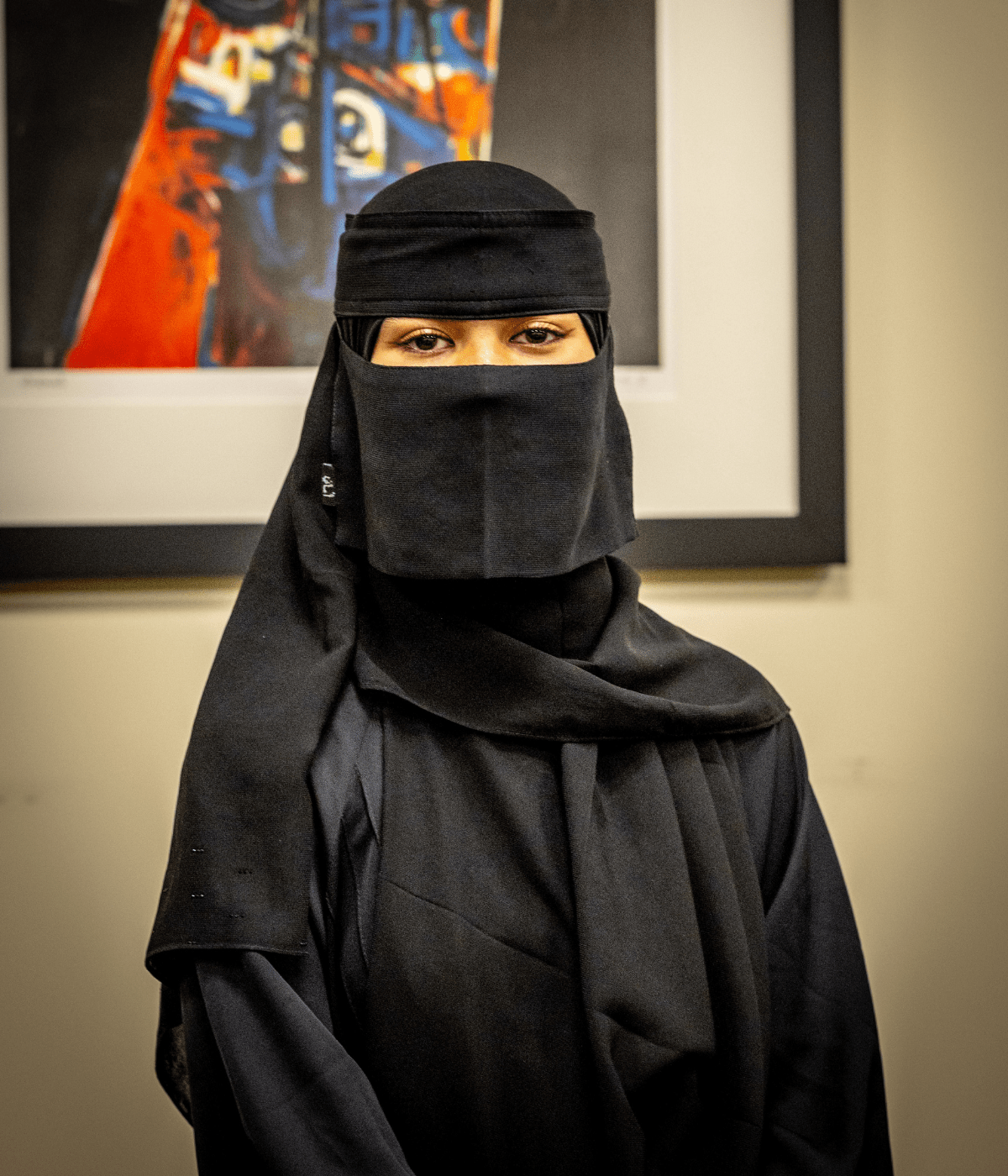 Portrait of a person wearing a black niqab and abaya, with only their eyes visible, standing indoors in front of a colorful abstract painting.