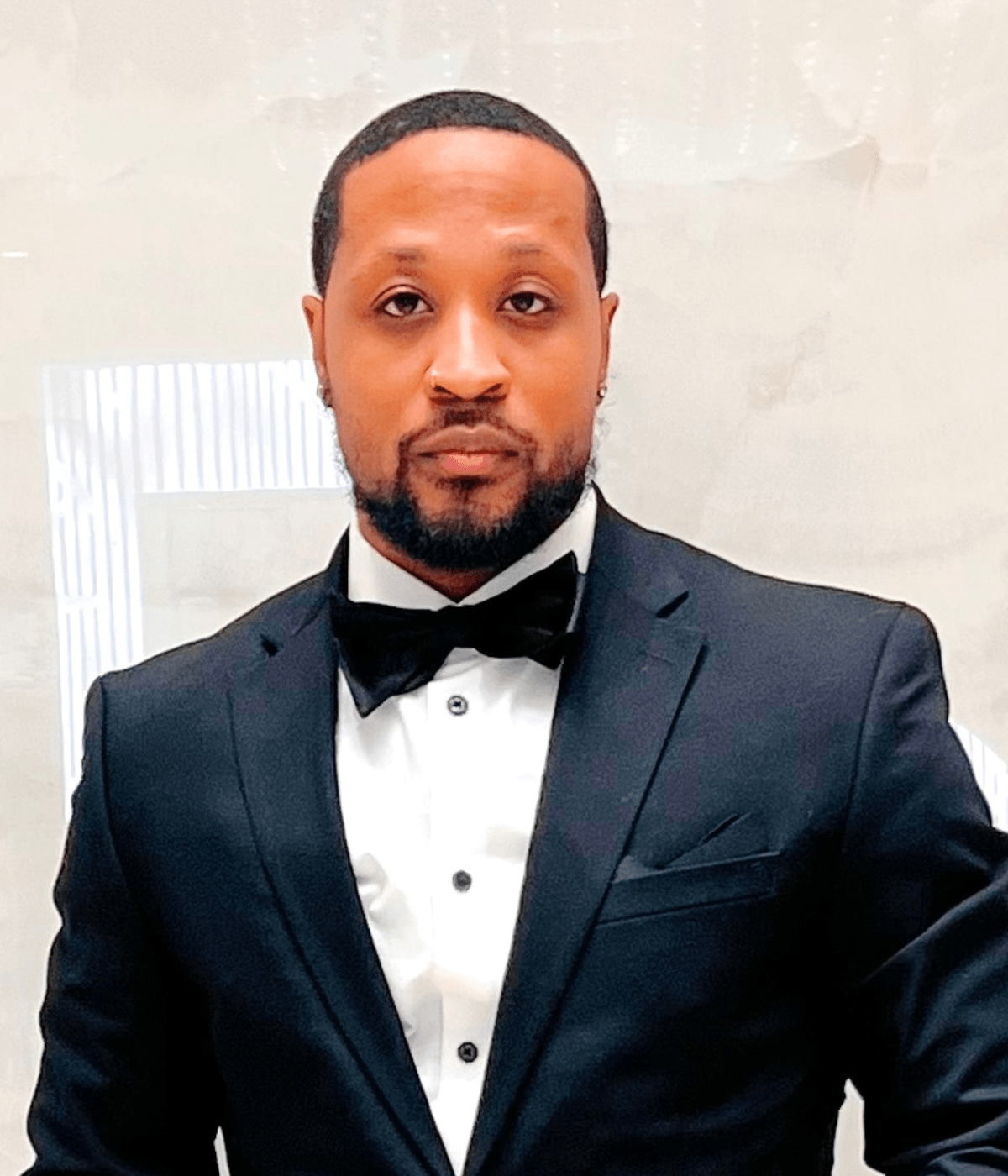 Portrait of a man in a black tuxedo and bow tie standing in front of a light, softly patterned background, looking directly at the camera with a neutral expression.