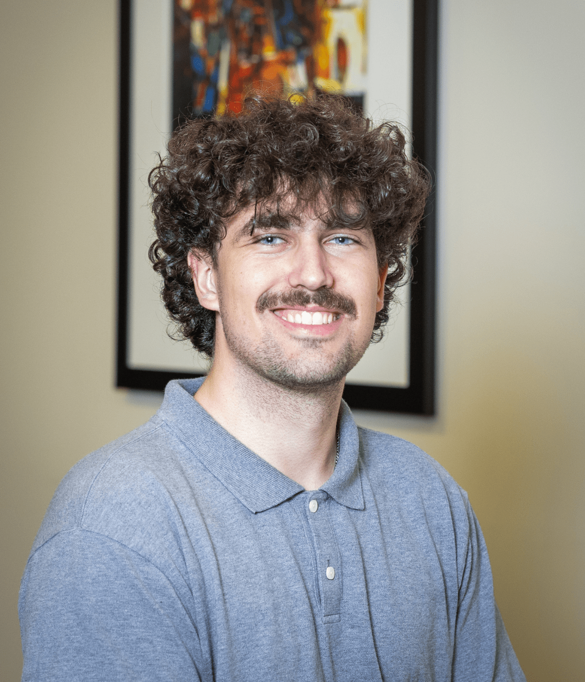 Portrait of a young man with curly brown hair and a mustache, smiling at the camera while wearing a light gray polo shirt, with a colorful abstract painting blurred in the background.